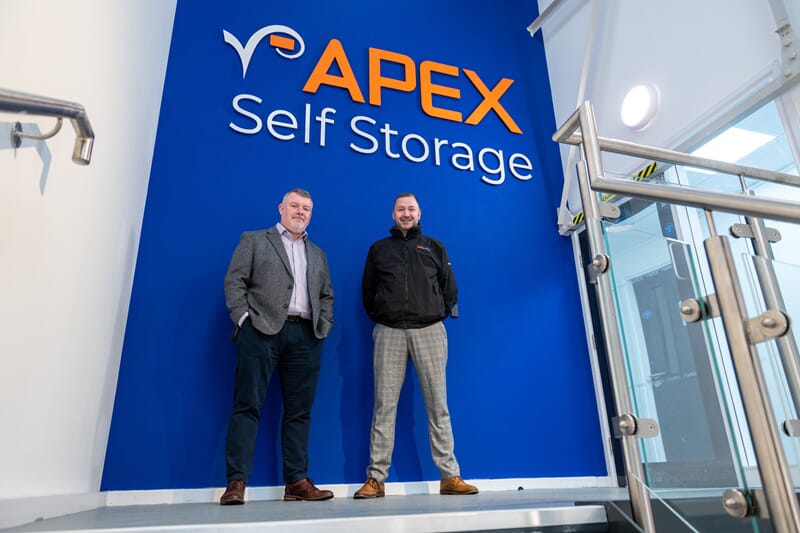 An image of two people in front of the Apex wall signage.